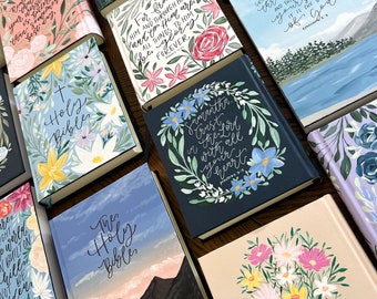 May include: A collection of personalized Holy Bibles with hand-painted floral designs and inspirational quotes. The covers feature a variety of floral patterns, including roses, lilies, and daisies, in shades of pink, blue, and yellow. Some covers also include mountain landscapes and sky scenes.