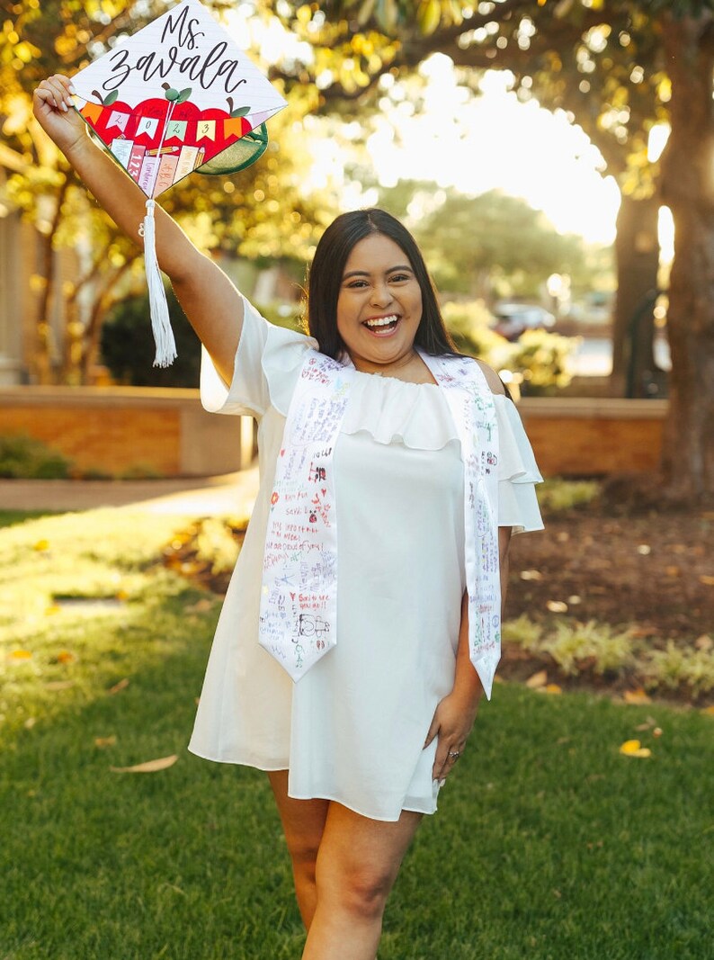May include: A white graduation cap decorated with a red apple graphic, the text Ms. Zavala, and the year 2023, featuring a white tassel. A white off-the-shoulder dress with ruffled sleeves and a white graduation stole covered in handwritten messages.