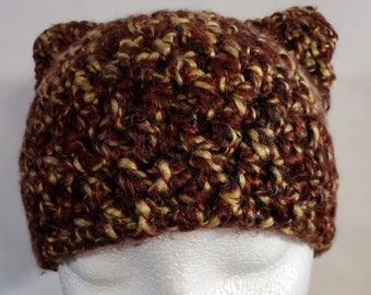 Puede incluir: Un gorro de punto de color marrón y dorado con orejeras. El gorro está hecho con un hilo texturizado y tiene un aspecto suave y acogedor.