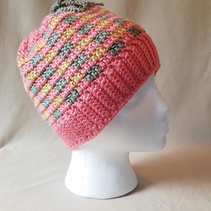 May include: A pink crocheted beanie with a green and yellow striped pattern. The beanie has a small tassel on top.