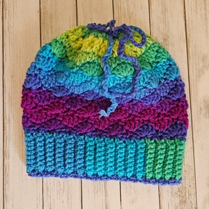May include: A colorful crocheted hat with a rainbow pattern in shades of green, blue, purple, and pink. The hat has a long crocheted tie that can be used to adjust the fit.