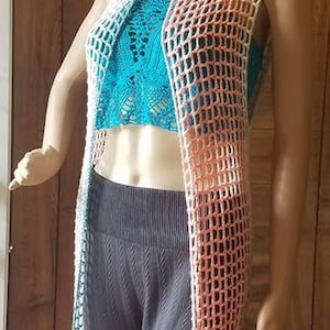 May include: A mannequin displays a crochet vest in shades of peach, white, and teal, layered over a turquoise top and dark gray pants. A matching headband completes the look. The open-weave vest is a long, sleeveless design.