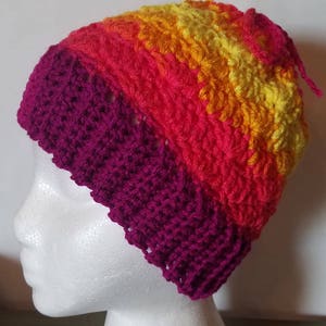 May include: A crocheted beanie hat with a multi-colored striped pattern in shades of yellow, orange, red, and pink. The hat has a solid pink crocheted brim.