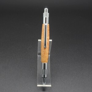 Full front view of click pen made with silk oak, a light brown wood with darker brown swirls in the grain, and chrome hardware. The pen is on a clear pen stand over a dark background.