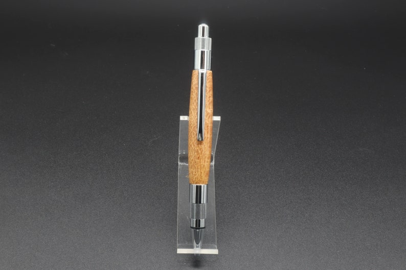 Full front view of click pen made with silk oak, a light brown wood with darker brown swirls in the grain, and chrome hardware. The pen is on a clear pen stand over a dark background.