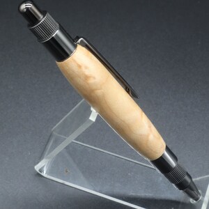 Close-up full left side view of click pen made with birds eye maple wood and black hardware. It's in a clear pen stand on a dark background. Maple wood is a light, cream-colored wood with darker brown swirls in the grain.