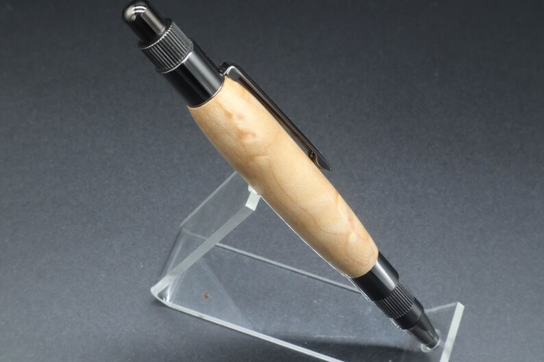 Close-up full left side view of click pen made with birds eye maple wood and black hardware. It's in a clear pen stand on a dark background. Maple wood is a light, cream-colored wood with darker brown swirls in the grain.