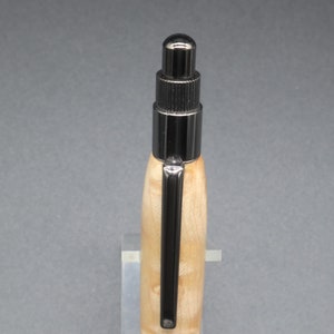 Close-up view of top of click pen made with birds eye maple wood and black hardware. It's in a clear pen stand on a dark background. Maple wood is a light, cream-colored wood with darker brown swirls in the grain.