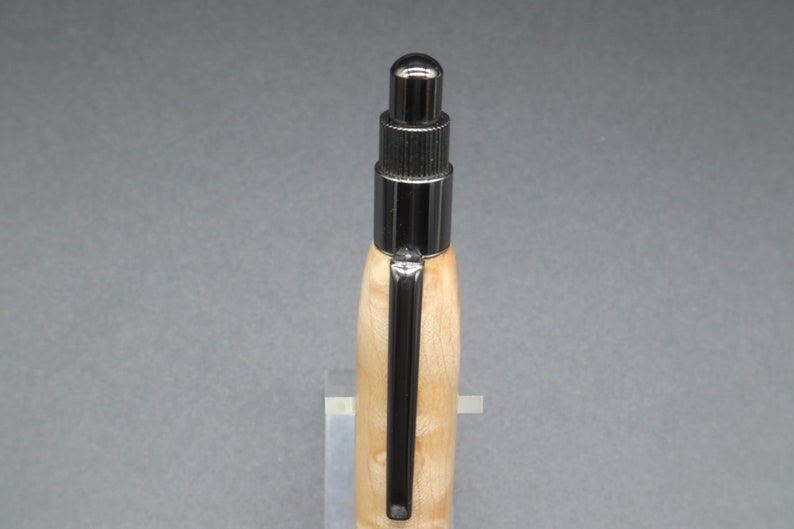 Close-up view of top of click pen made with birds eye maple wood and black hardware. It's in a clear pen stand on a dark background. Maple wood is a light, cream-colored wood with darker brown swirls in the grain.