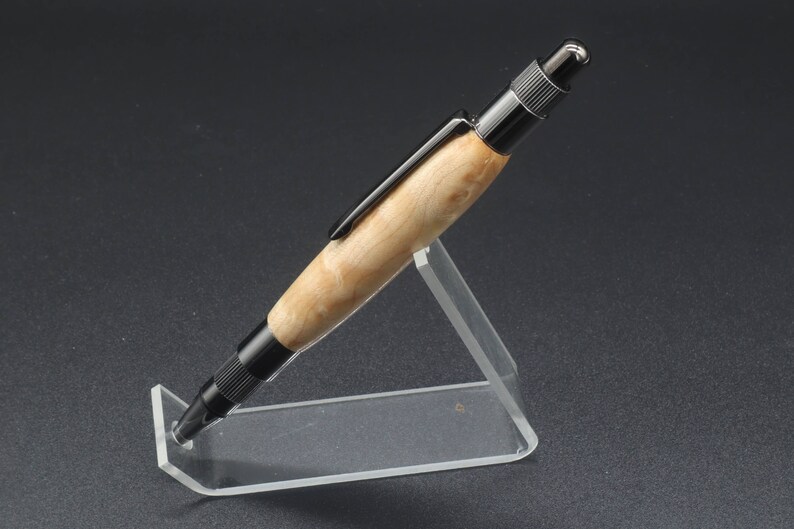 Full left side view of click pen made with birds eye maple wood and black hardware. It's in a clear pen stand on a dark background. Maple wood is a light, cream-colored wood with darker brown swirls in the grain.