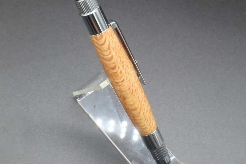 Close-up view of barrel of click pen made with silk oak, a light brown wood with darker brown swirls in the grain, and chrome hardware. The pen is on a clear pen stand over a dark background.