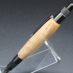 Close-up right side view of click pen made with birds eye maple wood and black hardware. It's in a clear pen stand on a dark background. Maple wood is a light, cream-colored wood with darker brown swirls in the grain.