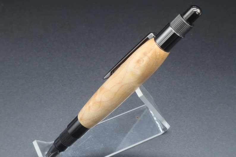 Close-up right side view of click pen made with birds eye maple wood and black hardware. It's in a clear pen stand on a dark background. Maple wood is a light, cream-colored wood with darker brown swirls in the grain.