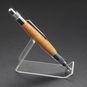 Full right side view of click pen made with silk oak, a light brown wood with darker brown swirls in the grain, and chrome hardware. The pen is on a clear pen stand over a dark background.