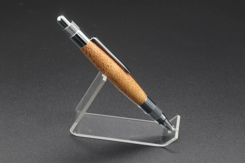 Full right side view of click pen made with silk oak, a light brown wood with darker brown swirls in the grain, and chrome hardware. The pen is on a clear pen stand over a dark background.