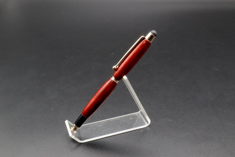 Side view of Bloodwood stylus pen with gold hardware on clear pen stand over black background