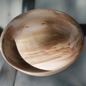 May include: A handcrafted wooden bowl with a natural, light brown finish. The bowl has a smooth, rounded shape and visible wood grain patterns. The interior shows variations in colour, with lighter and darker streaks. This decorative bowl is suitable for serving or display.