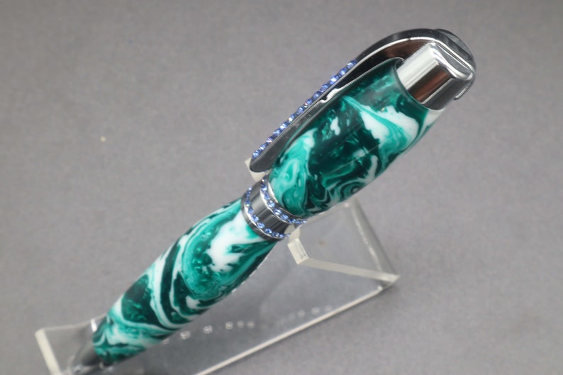 Close-up view of top of green and white (aka seafoam green) princess pen with blue crystals and chrome hardware on clear pen stand.