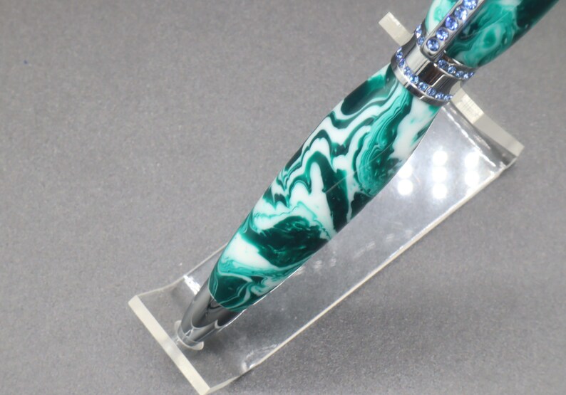 Close-up view of bottom/tip of green and white (aka seafoam green) princess pen with blue crystals and chrome hardware on clear pen stand.