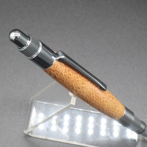 Close-up right side view of click pen made with silk oak, a light brown wood with darker brown swirls in the grain, and chrome hardware. The pen is on a clear pen stand over a dark background.