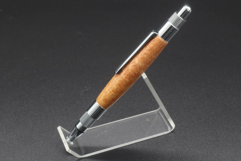 Full left side view of click pen made with silk oak, a light brown wood with darker brown swirls in the grain, and chrome hardware. The pen is on a clear pen stand over a dark background.