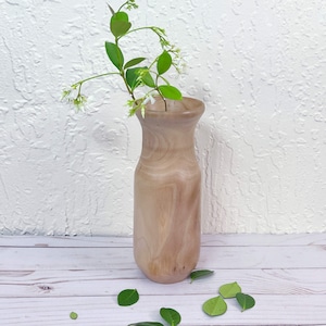 May include: A light brown wooden vase with a rounded base and a narrow neck, holding a sprig of white flowers with green leaves.
