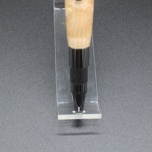 Close-up view of bottom of click pen made with birds eye maple wood and black hardware. It's in a clear pen stand on a dark background. Maple wood is a light, cream-colored wood with darker brown swirls in the grain.