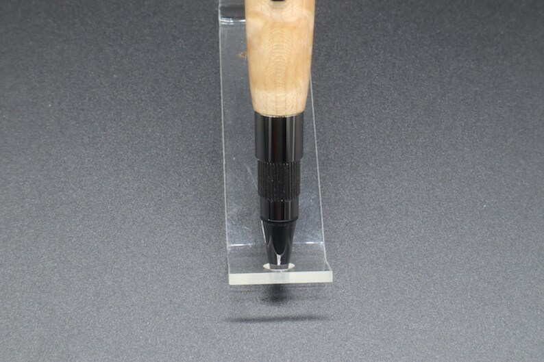 Close-up view of bottom of click pen made with birds eye maple wood and black hardware. It's in a clear pen stand on a dark background. Maple wood is a light, cream-colored wood with darker brown swirls in the grain.