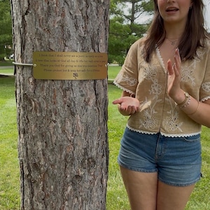 May include: A rectangular gold-colored metal plaque attached to a tree trunk with a silver wire. The plaque features black engraved text that reads: I think that I shall never see a poem as lovely as a tree. A tree that looks at God all day & lifts her leafy arms to pray. Thank you God for giving us this tree-mendous, Tree! Please protect her & keep her safe for a lifetime.