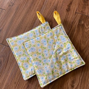 May include: Two yellow and white floral pot holders with yellow fabric loops. The pot holders are made of quilted fabric with a daisy pattern.