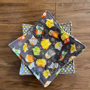 May include: Two square quilted bowl cozies. The top cozy features a dark gray background with colorful owl designs in yellow, orange, green, and gray. The bottom cozy has a gray polka dot pattern with yellow dots.