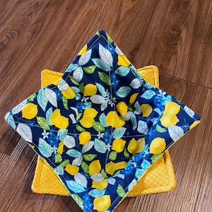May include: A set of two blue and yellow fabric pot holders with a lemon print. The pot holders are quilted and have a square shape.