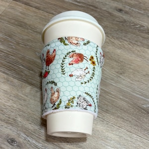 May include: A reusable cup sleeve featuring a light blue background with a white chicken wire pattern, decorated with illustrations of various chickens in shades of brown, white, and black, accented by small green leafy branches and yellow flowers.
