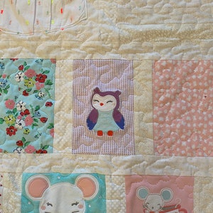 May include: A white quilt with a variety of fabric squares. The squares feature a floral pattern, a pink and white polka dot pattern, a purple and white checkered pattern, and an owl applique. There are also two squares with a light blue background and a white mouse applique.