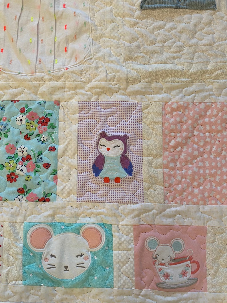 May include: A white quilt with a variety of fabric squares. The squares feature a floral pattern, a pink and white polka dot pattern, a purple and white checkered pattern, and an owl applique. There are also two squares with a light blue background and a white mouse applique.