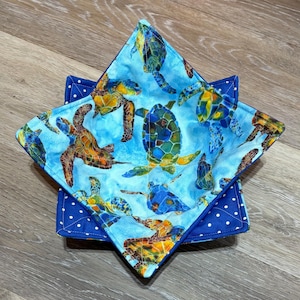 May include: Two square fabric bowl cozies. The top cozy features a blue background with colorful sea turtles. The bottom cozy is a solid blue with white polka dots. The cozies are designed to hold bowls and protect hands from heat.