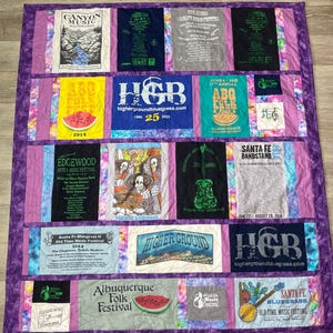 May include: A purple quilt with various patches of t-shirts featuring music festivals. The patches include the names of festivals like "Canyon Music", "Edgewood Arts & Music Festival", "Santa Fe Bandstand", "Higher Ground Bluegrass", "Albuquerque Folk Festival", and "Santa Fe Bluegrass Old Time Music Festival".