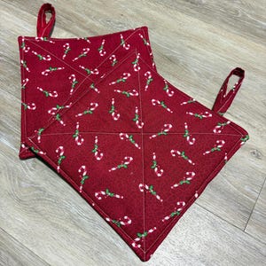 May include: Red quilted pot holders with a repeating pattern of white and green candy canes. The pot holders have a square shape with a loop for hanging. The fabric has a textured appearance.