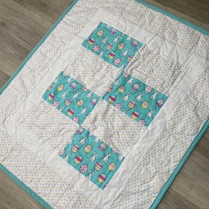 May include: A white quilt with a turquoise border and a patchwork design of turquoise squares with white bunnies and colorful Easter eggs. The quilt is quilted with a small white dot pattern.