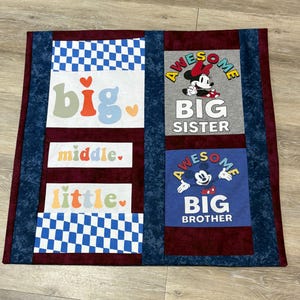 May include: A handmade quilt with a burgundy center and blue checkered and patterned borders. The quilt features the words "big", "middle", and "little" in colorful letters, along with images of Minnie and Mickey Mouse with the words "Big Sister" and "Big Brother".