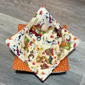 May include: A square, quilted bowl cozy with a cream-colored interior featuring a pattern of dogs, pumpkins, and fall leaves. The exterior is orange with white polka dots. The cozy is designed to hold a bowl.