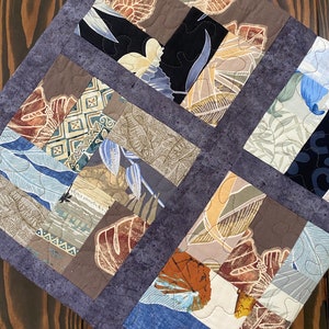 May include: A patchwork quilt with a variety of brown, blue, and beige fabrics featuring a tropical leaf pattern. The quilt is quilted with a white thread and has a dark gray border.