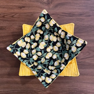 May include: A black and yellow bowl with a lemon print. The bowl is made of fabric and is designed to be used as a hot pad or trivet.