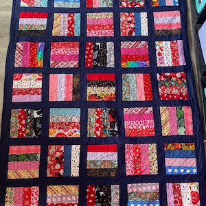 May include: A navy blue quilt with a patchwork design. The quilt is made up of squares of different fabrics, each with a different pattern. The fabrics are mostly red, pink, and white, with some black and gray accents.