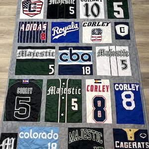 May include: A handmade quilt featuring patches of baseball jerseys and team logos. The quilt showcases various team names, numbers, and logos in a grid pattern, with colors including blue, green, white, and black. The quilt is on a gray background.