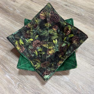 May include: A square fabric bowl cozy featuring a dark green background with a repeating pattern of brown pinecones and green pine needles. The interior is quilted with white thread in a cross pattern, and the exterior is a solid dark green fabric.