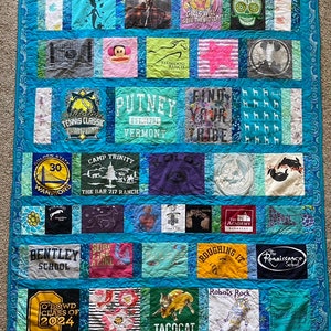 May include: A blue and white quilt made from t-shirts with various logos and designs. The quilt is made up of squares and rectangles, and the designs include a basketball team logo, a school logo, a camp logo, a horse, a monkey, a skull, a mermaid, a girl with a star, a cat, a tree, a heart, a fish, a robot, a raccoon, a butterfly, a taco, and more. The quilt is surrounded by a blue border.