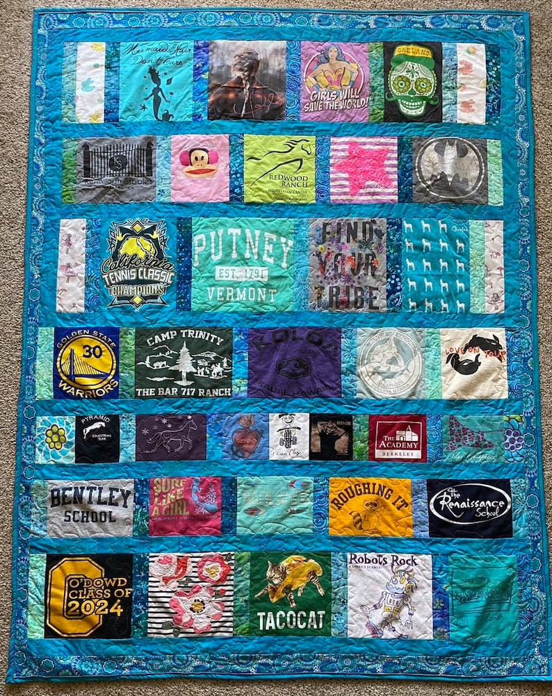 May include: A blue and white quilt made from t-shirts with various logos and designs. The quilt is made up of squares and rectangles, and the designs include a basketball team logo, a school logo, a camp logo, a horse, a monkey, a skull, a mermaid, a girl with a star, a cat, a tree, a heart, a fish, a robot, a raccoon, a butterfly, a taco, and more. The quilt is surrounded by a blue border.