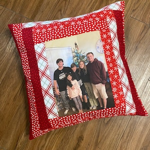 May include: A red and white Christmas pillow with a photo of a family in front of a Christmas tree. The pillow has a plaid border and a white snowflake pattern.