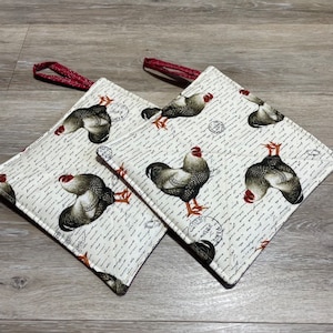 May include: Two white fabric pot holders with red hanging loops. The pot holders feature a repeating pattern of black and white roosters on a background of handwritten script.